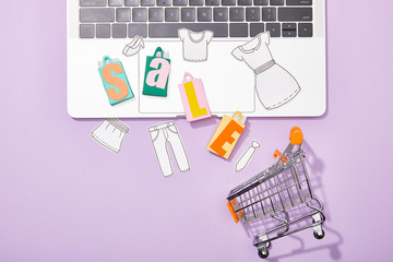 top view of colorful toy bags with sale lettering and paper cut of clothes on laptop falling in toy shopping cart on violet