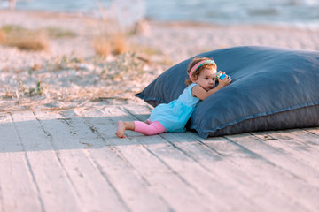 Beautiful baby in a turquoise dress on the beach is resting on a large pillow.