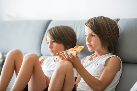 Cute children, sitting on couch, eating pizza and watching TV. Hungry child taking a bite from pizza on a pizza party