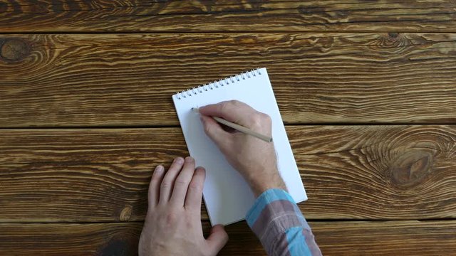 The man is writing in a notebook. He tries to make a list of ideas and puts a question mark. On a wooden table. 4K 2160p 25fps UHD video.