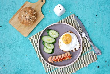 Breakfast, fried egg with sausage and slices of fresh cucumbers on a brown clay plate on a turquoise concrete background. Children's breakfasts. School breakfast concept. Top view, copy space.