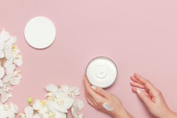 Young woman moisturizes her hand with cosmetic cream lotion opened container with cream body milk White Phalaenopsis orchid flowers on pink background Flat lay top view minimalism style Beauty concept