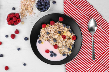 black bowl of oat granola, muesli with yogurt, fresh raspberries, blueberries on white wooden board for healthy breakfast. top view