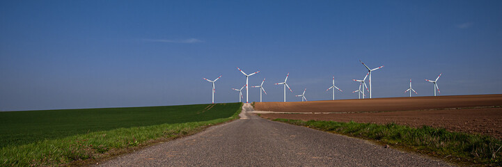 Fototapeta premium Wind farm in front of a blue sky, green grass to the left and brown field with brown earth to the right of the dirt road
