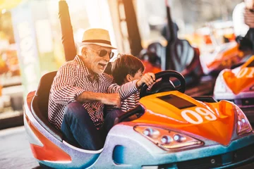 Fotobehang Grootvader en kleinzoon hebben plezier en brengen goede quality time samen door in het pretpark. Ze genieten en glimlachen terwijl ze samen in een botsauto rijden. © Dusko