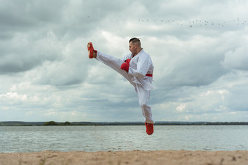 A man in a sports kimono with a red belt fulfills a karate jump kick. Training takes place on the beach. against the background of the sea.