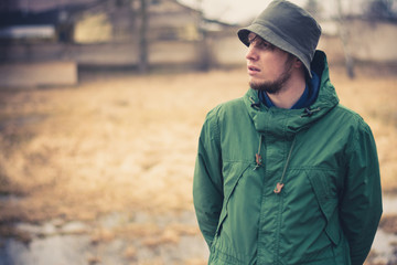 a young guy in a swampy area stares away. Casual wear, khaki pants, green jacket, green panama. In the background a swamp and flooded houses © Anton