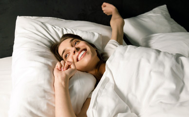 Fototapeta na wymiar Beautiful brunette woman waking up in her white bed, she is smiling and stretching