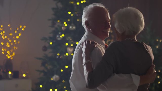 Handheld medium shot of happy senior man and woman with sparkler dancing in dark room with lit Christmas tree