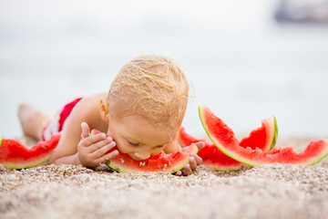 Cute toddler boy, blond child, eating watermelon on the beach coast
