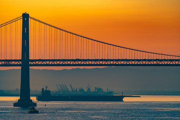 Foggy/Misty summer morning sunrise in California and silhouette of the iconic Oakland Bay Bridge in San Francisco as cargo ship sails by. Famous travel location landmark in the west coast city.