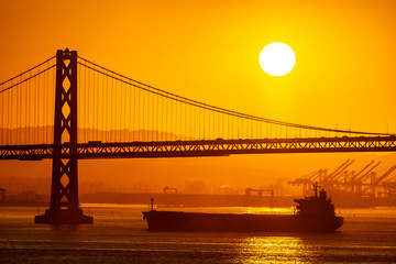 Fototapeta na wymiar Foggy/Misty summer morning sunrise in California and silhouette of the iconic Oakland Bay Bridge in San Francisco as cargo ship sails by. Famous travel location landmark in the west coast city.