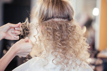 Rear view of the hands of a hairdresser master makes a curly hairstyle for a girl client a blonde woman before preparing for the holiday event. The concept of creating a spectacular hairstyle