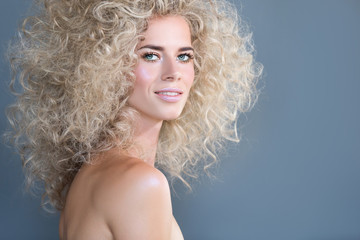 Close-up portrait of a smiling cheerful curly young blonde girl with bright art makeup posing on a gray background. Magazine glamor concept