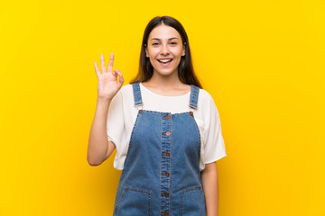 Young woman in dungarees over isolated yellow background surprised and showing ok sign