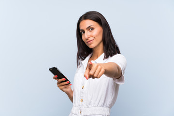 Young woman with a mobile phone over isolated blue wall points finger at you with a confident...
