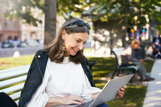 People, age, lifestyle, technology and communication. Outdoor image of beautiful retired female dressed in stylish clothes sitting on bench with touch pad pc on her lap, watching funny video, laughing