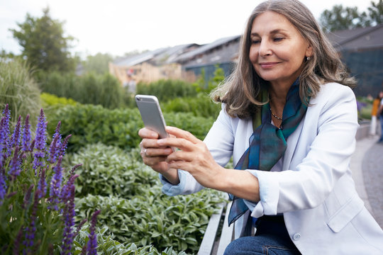 Elegant middle aged European female holding smart phone, trying to capture blossom of violet flowers, walking in park. Smiling mature woman taking picture of beautiful nature around her using mobile