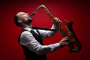 Naklejka na ściany i meble Portrait of professional musician saxophonist man in suit plays jazz music on saxophone, red background in a photo studio