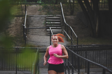 Beautiful adult red haired woman athlete in pink top and shorts makes amorning run on the city...