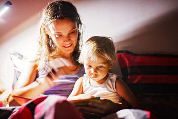 Obraz premium Mother, reading a book to her toddler boy at home at night, night stand lamp turned on