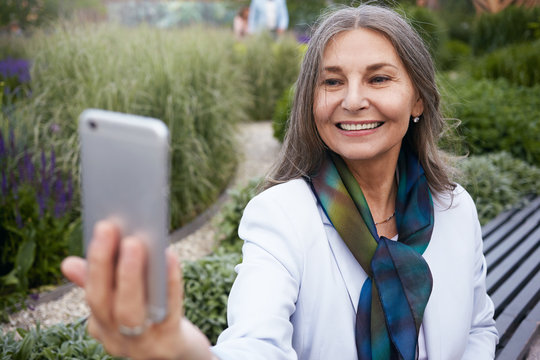 Horizontal image of beautiful positive Caucasian woman pensioner taking selfie using mobile phone, looking with broad joyful smile, spending leisure time outdoors. Selective focus on electronic gadget