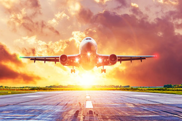 Airplane lands on runway with landing lights on during a bright rays sunset, with beautiful clouds. - obrazy, fototapety, plakaty