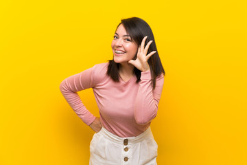 Young Mexican woman over isolated yellow background listening to something by putting hand on the ear