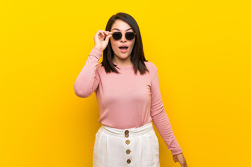 Young Mexican woman over isolated yellow background with glasses and surprised