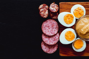 Healthy and fresh organic food served as appetizer with copy space - Hardboiled egg halves and slices of smoked salami and sausage on black stone textured background