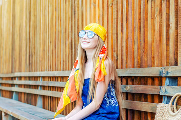 Young girl in a yellow bandana and large sunglasses portrait on the background of a wooden wall of red boards. Sky, clouds reflected in glasses. Summer rest.