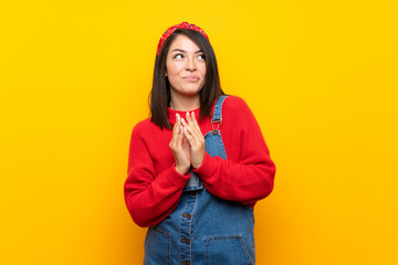 Young Mexican woman with overalls over yellow wall scheming something