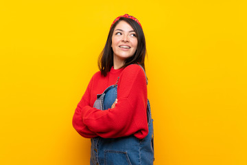 Young Mexican woman with overalls over yellow wall with arms crossed and happy
