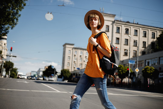 Young girl with headphones in a yellow t-shirt and a straw hat  crossing the road with a backpack in a city street on a summer sunny day