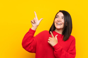 Young Mexican woman with red sweater over yellow wall pointing with the index finger a great idea