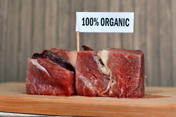 Big raw red meat chunks on wooden plate with label saying '100 percent organic', concept for healthy organic food production