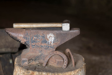 Hammer and anvil on a wooden block in the stable. Two used rusty horseshoes stand nearby. Dark background. The place for an inscription