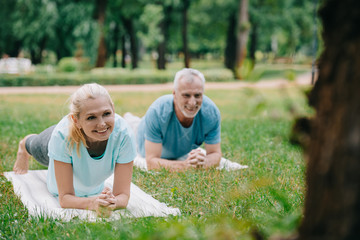 selective focus of smiling mature man and woman doing plank exercise while practicing yoga in park