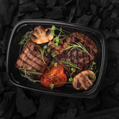 Grilled meat steak with vegetables in black plastic box to go