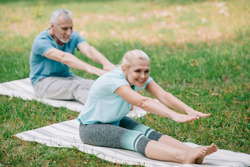 Naklejka na ściany i meble smiling mature man and woman practicing yoga poses while sitting on yoga mats