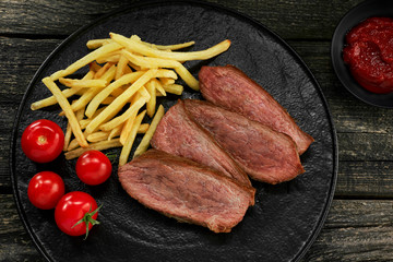 Sliced beef steak with French fries and cherry tomatoes on a black plate. Delicious American lunch. Top view, directly above shot.