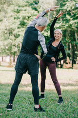 Fototapeta premium back view of mature sportsman warming up near attractive, smiling sportswoman in park
