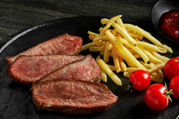Steak with French fries, fresh cherry tomatoes and sauce on a rustic wooden table. Wholesome classic American lunch on a black plate. Closeup shot.