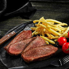 Sliced beef steak with French fries, sauce and cherry tomatoes on a black plate. Delicious American lunch with red wine ona rustic wooden table.