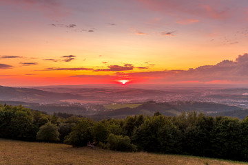 Sunset with view on landscape with fully colored clouds and orange sun goes down and city Valasske Mezirici captured during summer late time