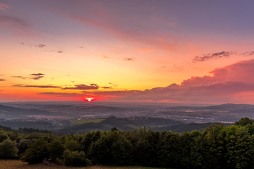 Naklejka na ściany i meble Sunset with view on landscape with fully colored clouds and orange sun goes down and city Valasske Mezirici captured during summer late time