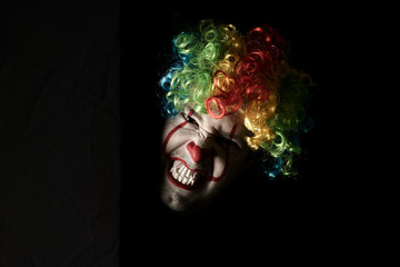 Close-up of a scary clown peeping around the corner of a black wall. He is wearing a colored wig and sharp fangs.