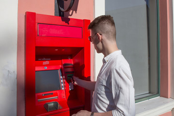 young man student withdrawing money from a bank cash point, outdoors