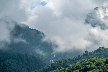 Green mountains with a high waterfall in fog and clouds.