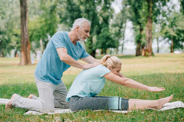 handsome mature man helping woman practicing yoga pose on lawn in park